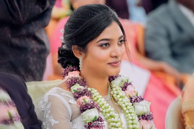 Bride in a lace wedding dress wearing pink and white flower garlands, jewelry, and soft makeup during a ceremony.