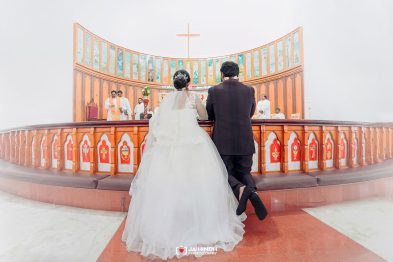 Bride in a white wedding gown and groom in a dark suit walk toward the church altar, viewed from behind inside a church.