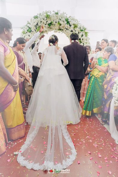 Bride in a white gown and veil walks under a white floral arch with a groom in a dark suit, surrounded by colorful guests at a traditional wedding.