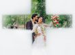 Couple in wedding attire embracing and smiling, adorned with flower garlands, seen through a cross-shaped opening amid greenery.