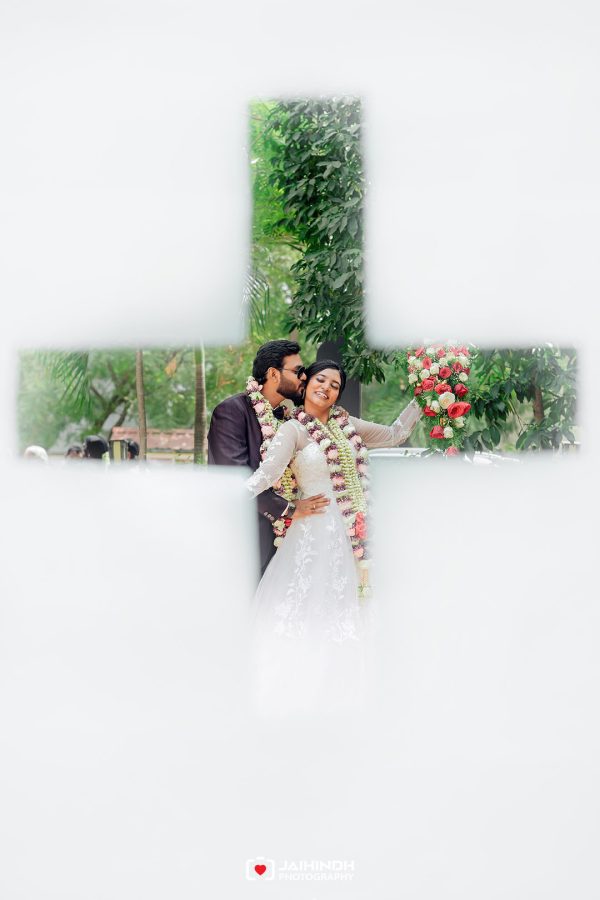 Best Wedding Photographers in Madurai 46 Couple in wedding attire embracing and smiling, adorned with flower garlands, seen through a cross-shaped opening amid greenery.