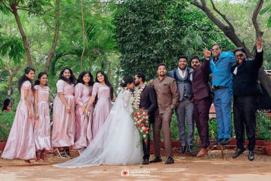 Bride and groom kissing center stage at an outdoor wedding, surrounded by bridesmaids in pink dresses and groomsmen in colorful suits, some jumping for joy under trees.