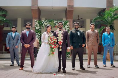 Bride and groom at center in wedding attire with garlands, flanked by groomsmen in colorful suits and guests, outdoors with floral arch behind them.