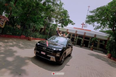 Couple in an open-top black SUV with a red ribbon, raising hands in celebration outside CSI Cathedral Madurai.