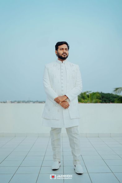 Full-body portrait of a man in a white ceremonial sherwani standing on a tiled terrace against a clear blue sky.