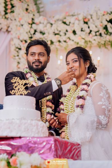 Newlywed couple feeding each other cake at their wedding, wearing floral garlands with a lush flower backdrop behind them.
