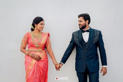Couple in wedding attire holding hands and looking at each other against a plain wall. - Bride in pink sari with gold jewelry and the groom in a navy suit.