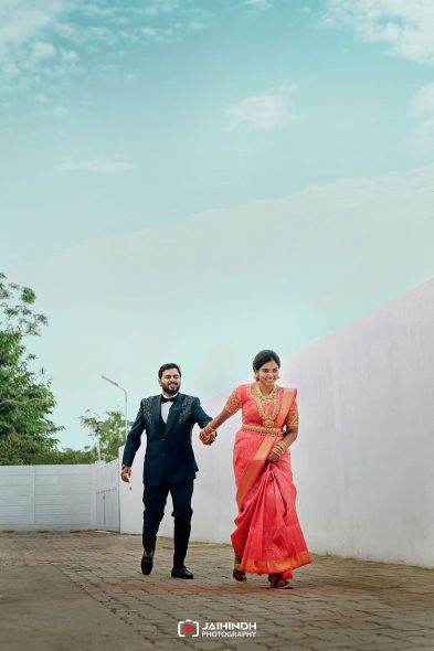Indo‑Indian bride in pink saree with gold jewelry walking hand in hand with a groom in a black suit along a paved path beside a white wall on a sunny day