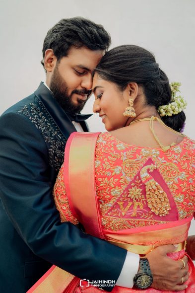 A couple embraces closely, man in a dark embroidered suit and bow tie, woman in a pink embroidered saree with gold jewelry and jasmine flowers in her hair.