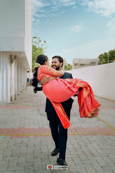 Man in a suit lifts a smiling woman in a bright pink traditional dress outdoors, courtyard setting, clear blue sky.