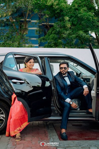 Couple in formal attire sit in a black car with doors open; woman in pink sari smiles, man in navy suit and sunglasses.