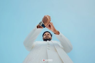 Low-angle shot of a bearded man in a white traditional outfit, hands joined overhead against a bright blue sky.