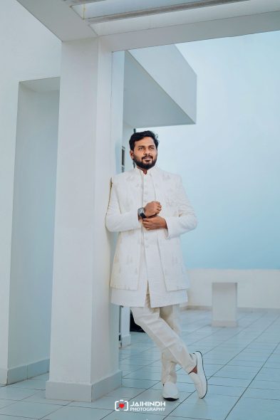 Man in a white sherwani posing against white architectural columns on a rooftop, looking at the camera.