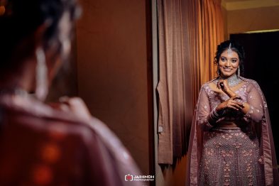 Bride in a pink embellished lehenga smiles at her reflection in a mirror by a window/curtain backdrop