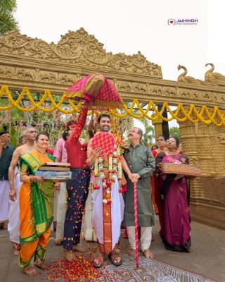Indian wedding ceremony under a gold carved arch, couple adorned with garlands, surrounded by family.