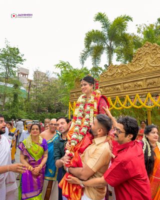 Bride in red and gold being carried on shoulders, adorned with garlands at an outdoor Indian wedding with a gold ornate backdrop.