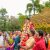Bride in red and gold being carried on shoulders, adorned with garlands at an outdoor Indian wedding with a gold ornate backdrop.