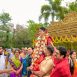 Bride in red and gold being carried on shoulders, adorned with garlands at an outdoor Indian wedding with a gold ornate backdrop.