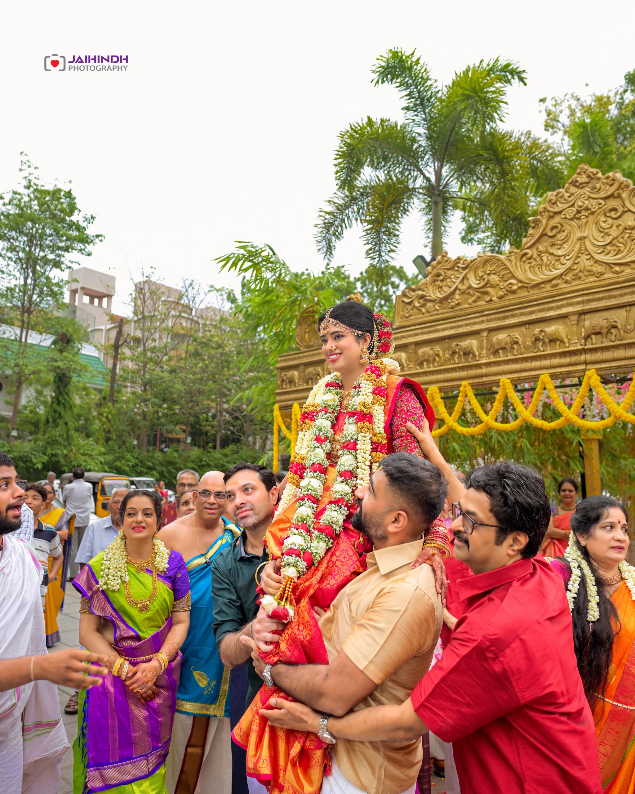 Bride in red and gold being carried on shoulders, adorned with garlands at an outdoor Indian wedding with a gold ornate backdrop.