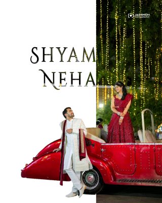 Bride and groom in traditional Indian attire pose beside a bright red vintage car under string lights at a wedding.